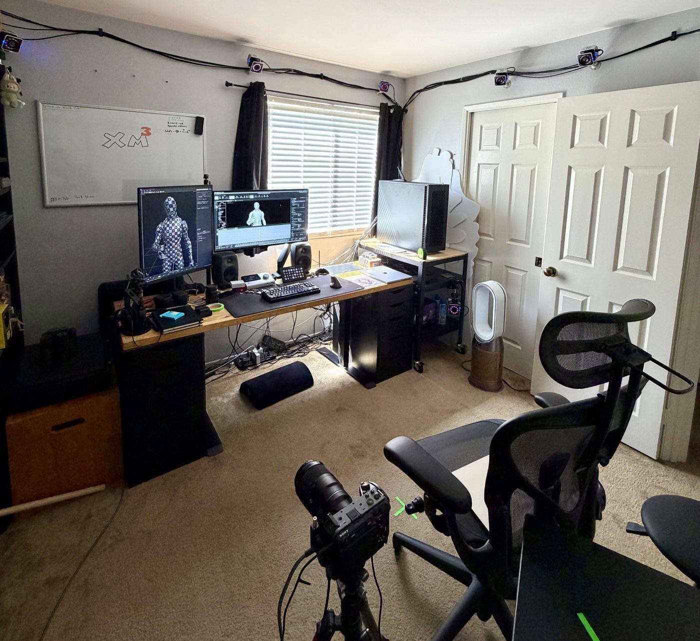 The original XM3 room: a desk with two monitors showing motion capture software, motion capture cameras mounted along the tops of the walls, and a camera on a tripod in the foreground.